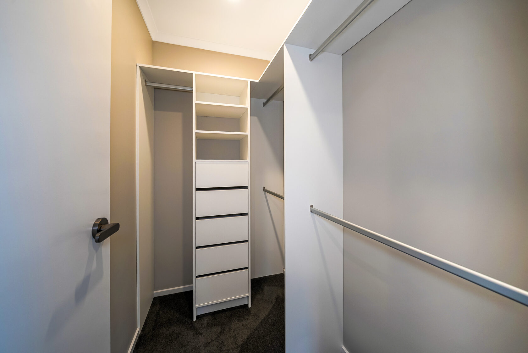 Walk-in wardrobe with built-in white shelving, four drawers, and hanging rails on both sides, finished with dark carpet and soft ceiling lighting.