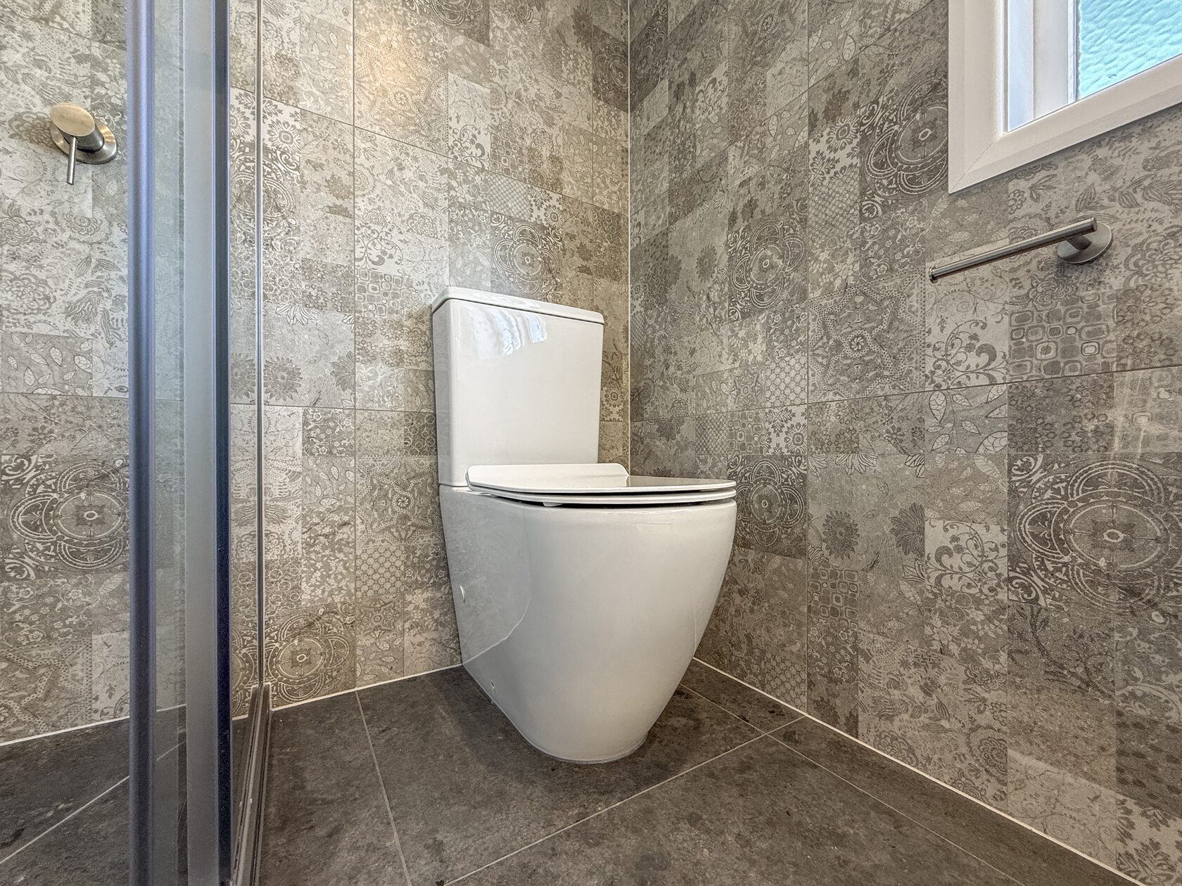 White toilet beside a glass shower in a bathroom with patterned grey tiles