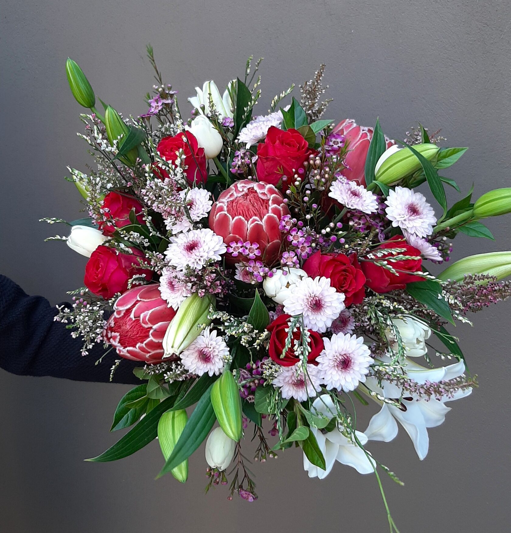 A luxurious and vibrant bouquet harmoniously combining large pink proteas, red roses, white and soft pink chrysanthemums, as well as white lilies and tulips.
