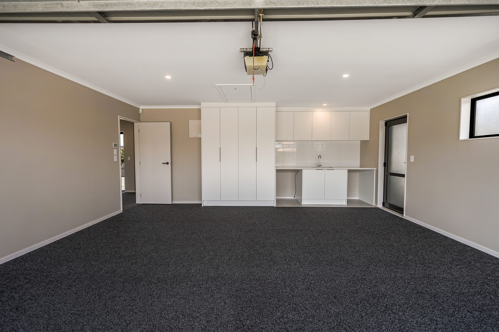 Spacious carpeted garage or utility room with built-in white storage cabinets, laundry sink area, neutral walls, and ceiling-mounted garage door opener.