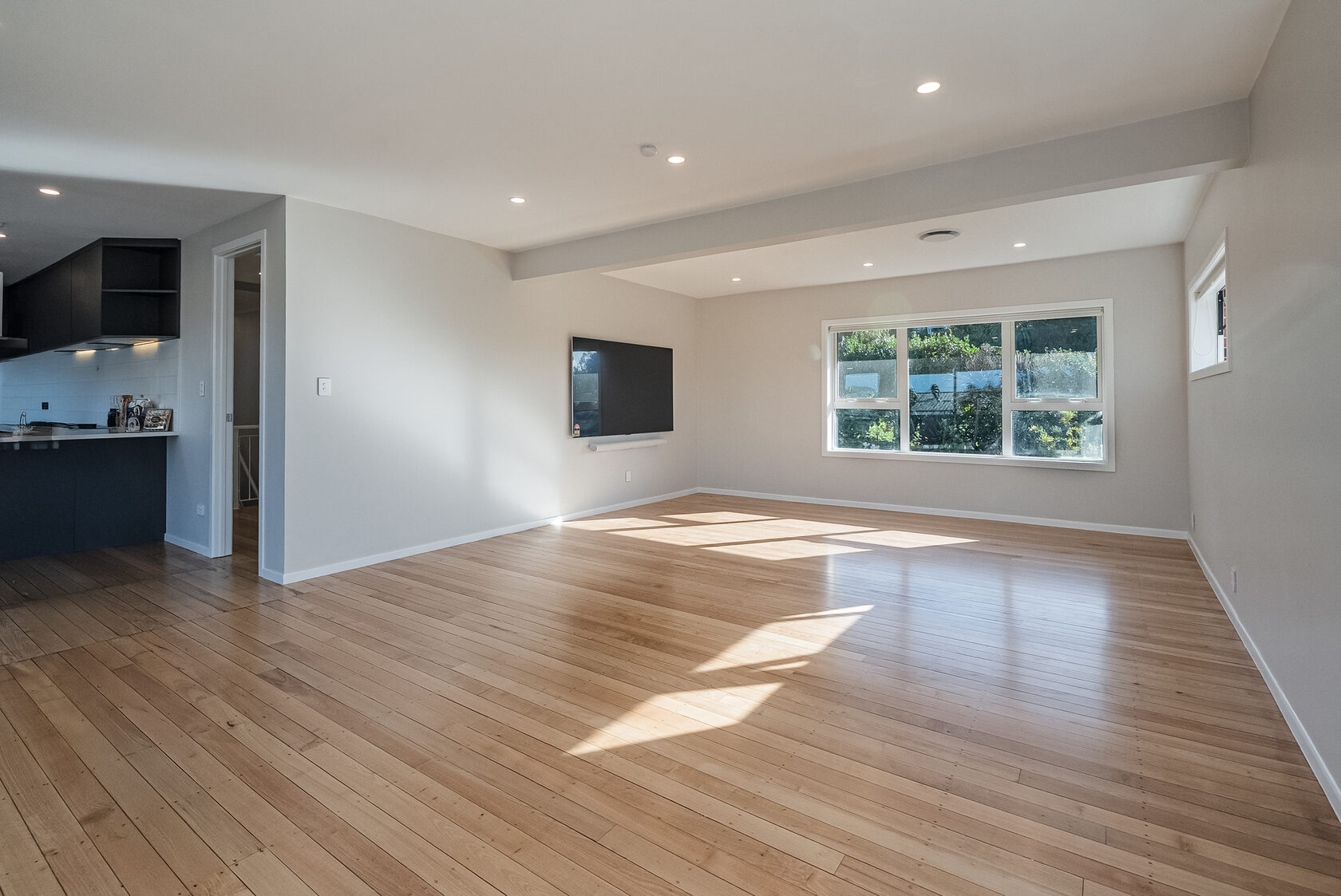 Wide view of the renovated Glenfield living area with the dark kitchen visible to the left, timber flooring and a large sunlit window