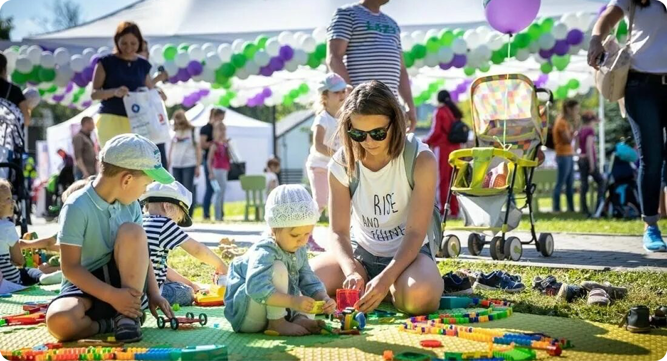 Svetao i sunčan Porodični dan (Porodični dan) na otvorenom. Mama i deca sede na zelenom leglu i strastveno se igraju sa svetlim, raznobojnim konstruktorima. U pozadini je svečana atmosfera sa belim šatorima, vijencima ljubičasto-bijelo-zelenih kuglica i šetajućim gostima. Odličan primer letnjeg porodičnog festivala ili korporativnog događaja za zaposlene sa decom.