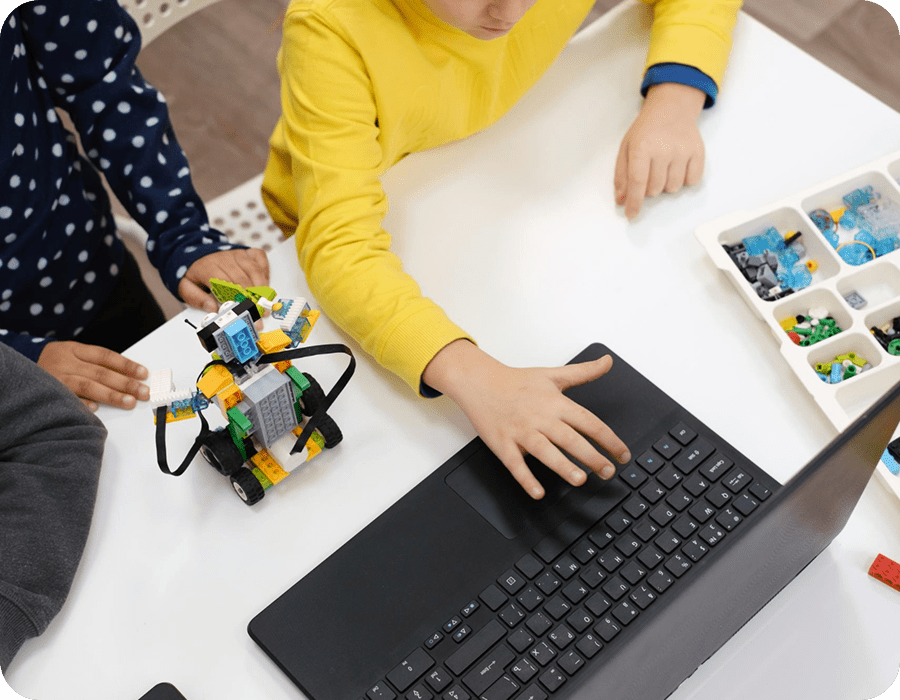 Seen from above: children around a white table with a small LEGO&reg; robot on wheels, a tray of LEGO&reg; parts and a laptop