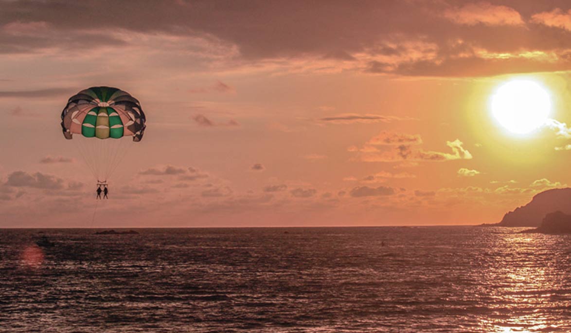 Dos personas en parasailing bajo un paracaídas verde y blanco sobre el mar al atardecer