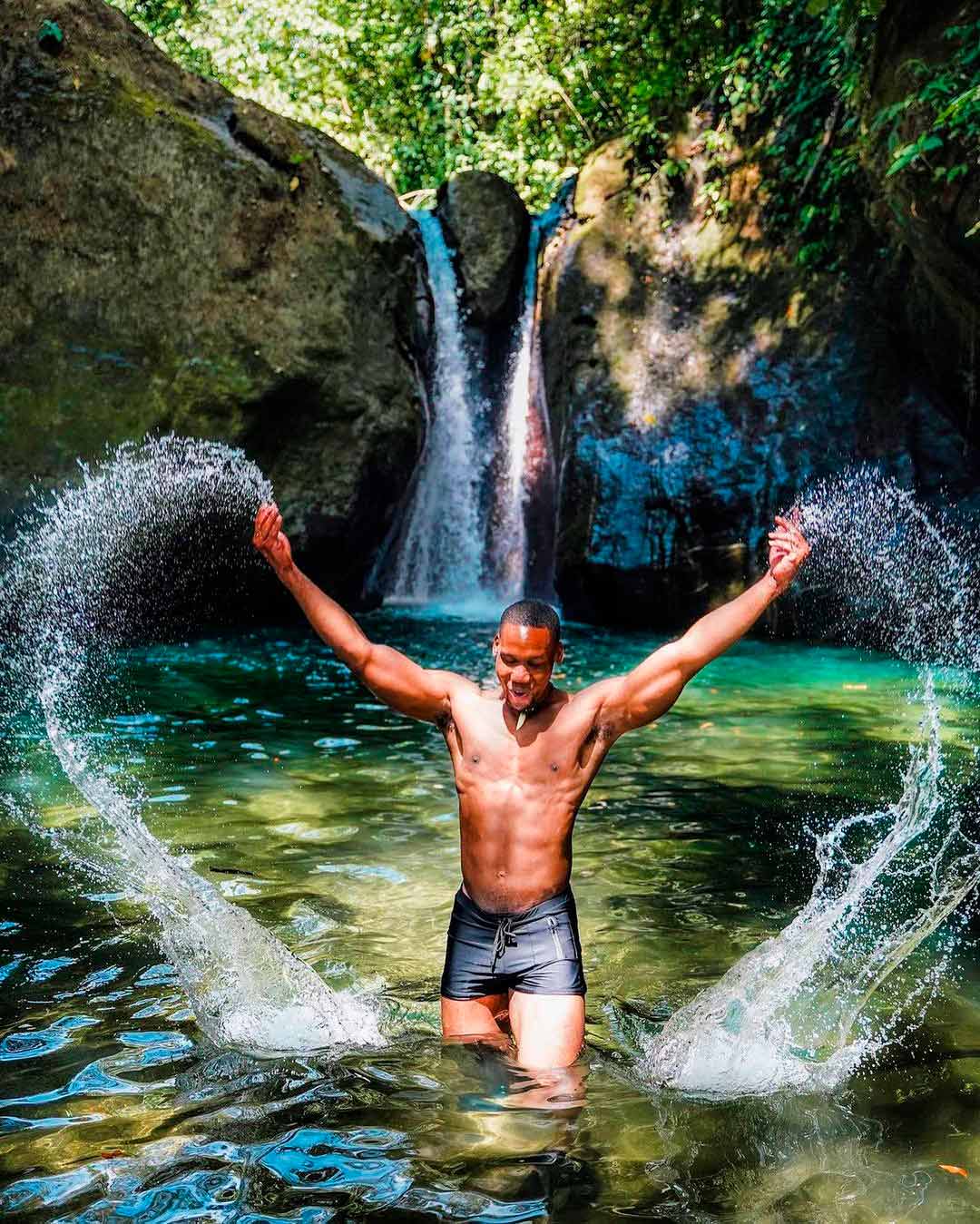 Hombre chapoteando en una poza natural frente a una pequeña catarata rodeada de roca y follaje