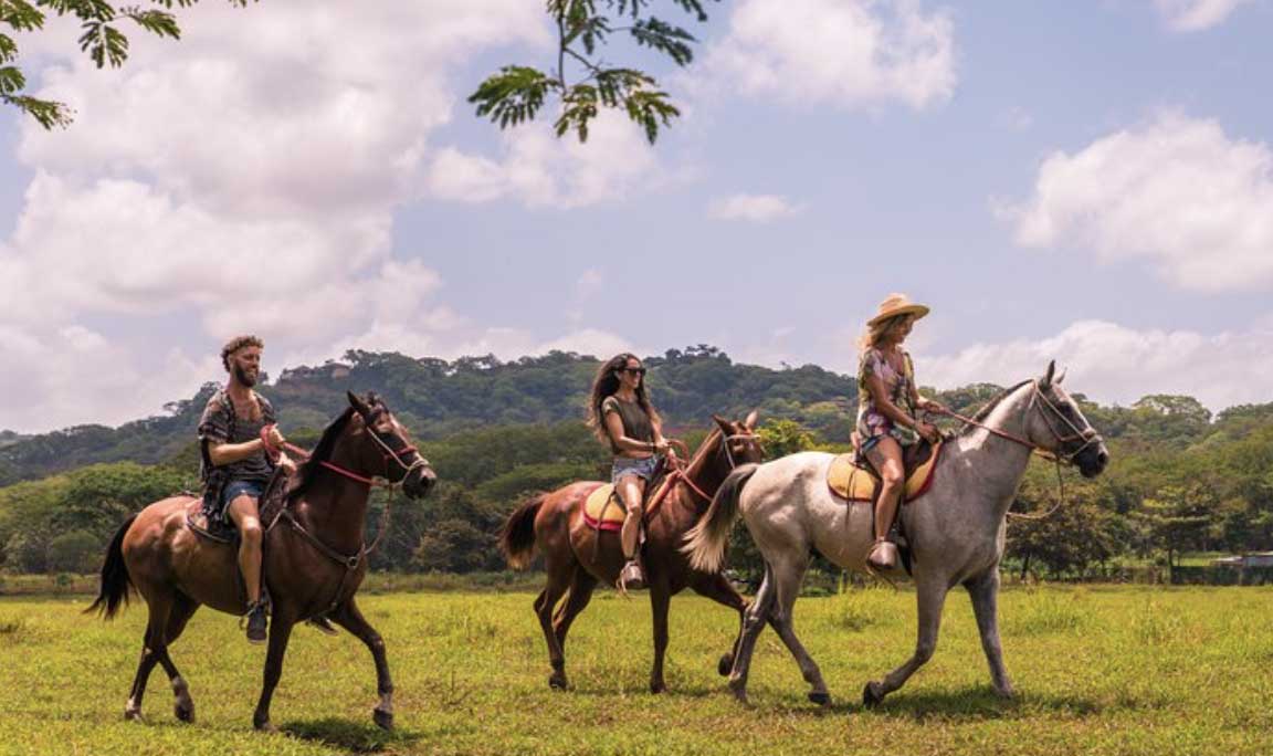 Tres jinetes a caballo cruzando un campo verde con árboles y colinas al fondo