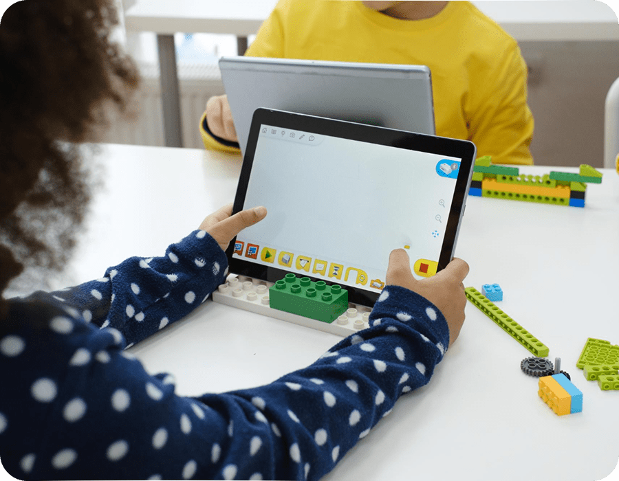A child in a polka-dot top using a tablet that rests on a green LEGO&reg; baseplate, with LEGO&reg; pieces on the table