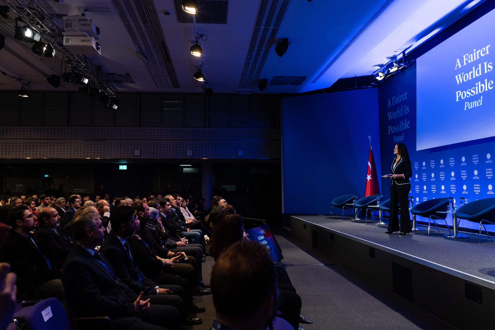 Wide view of a conference audience and a speaker on a blue-lit stage