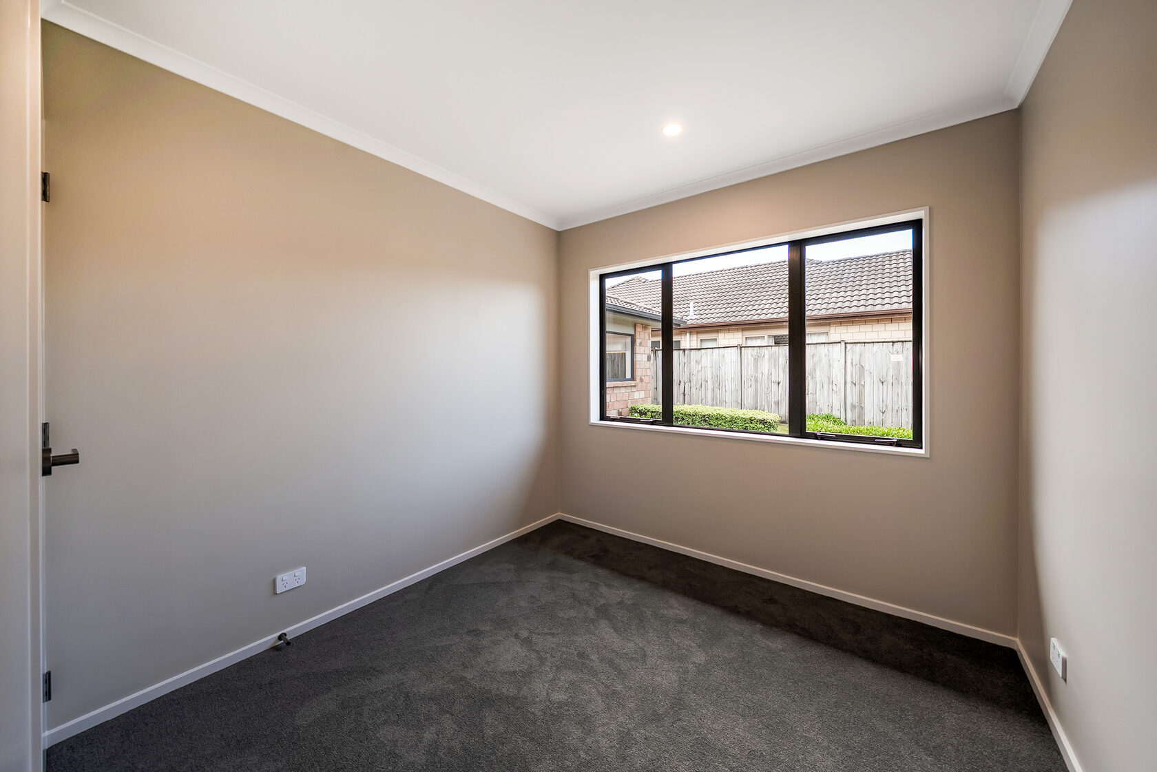 Flat Bush home renovation: Empty bedroom with neutral walls, dark carpet, and large window overlooking a fenced backyard.