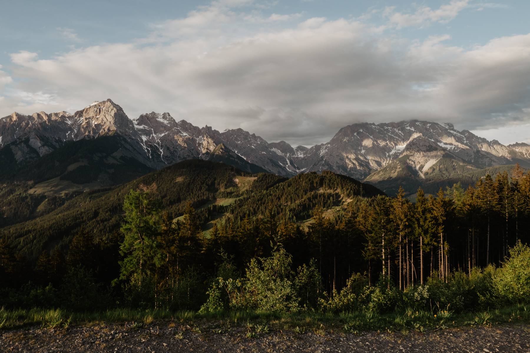 Jagged, snow-dusted alpine peaks rising above forested mountain slopes beneath a softly clouded sky