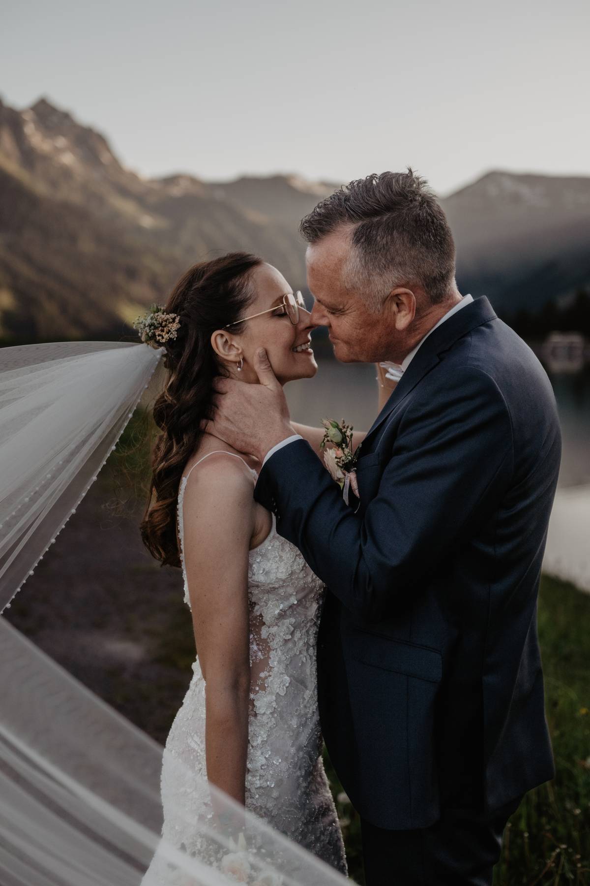 Mountain elopement couple kissing by an alpine lake at sunset, veil flowing with peaks behind