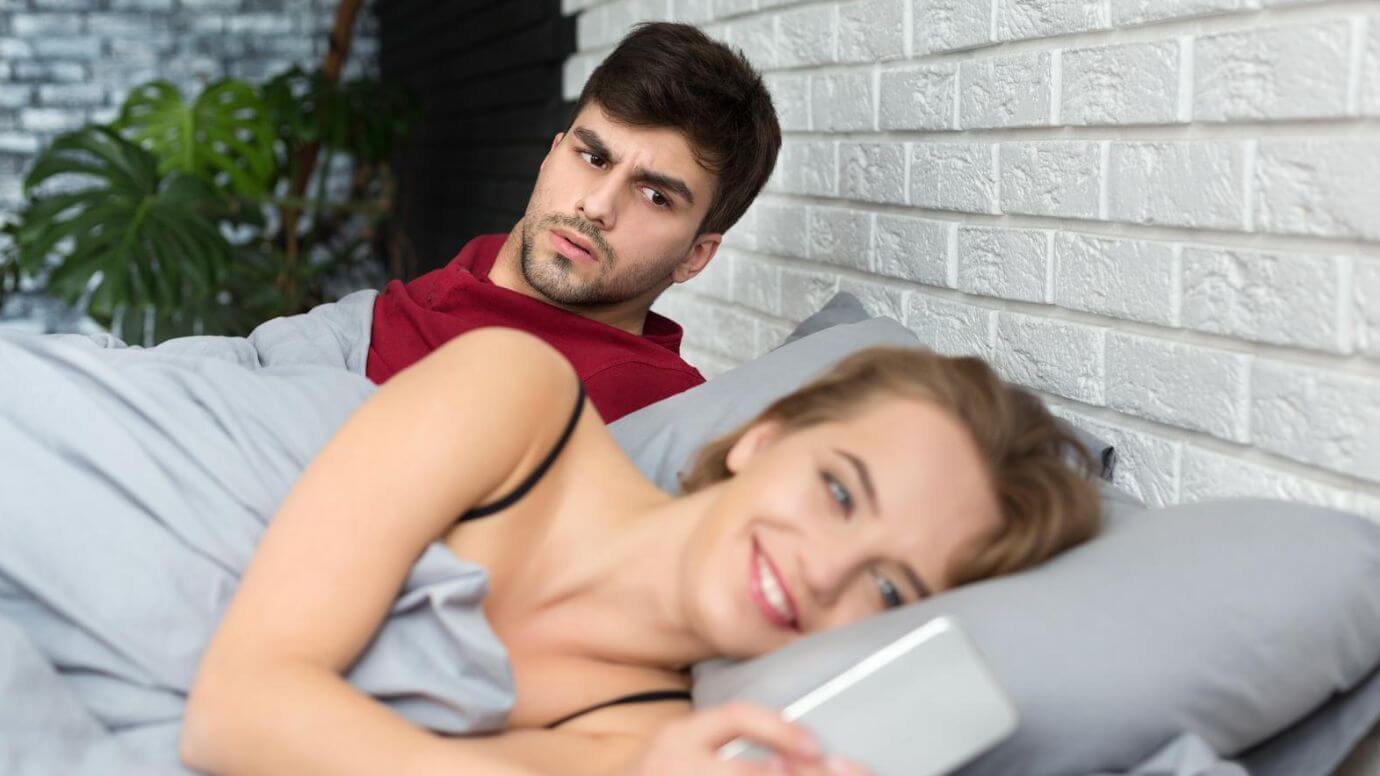 Upset man sitting on the edge of the bed with a seemingly oblivious woman lying behind him