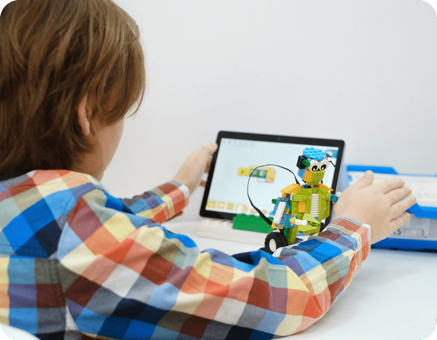 A child in a checked shirt holding a tablet next to a green LEGO&reg; robot they have built