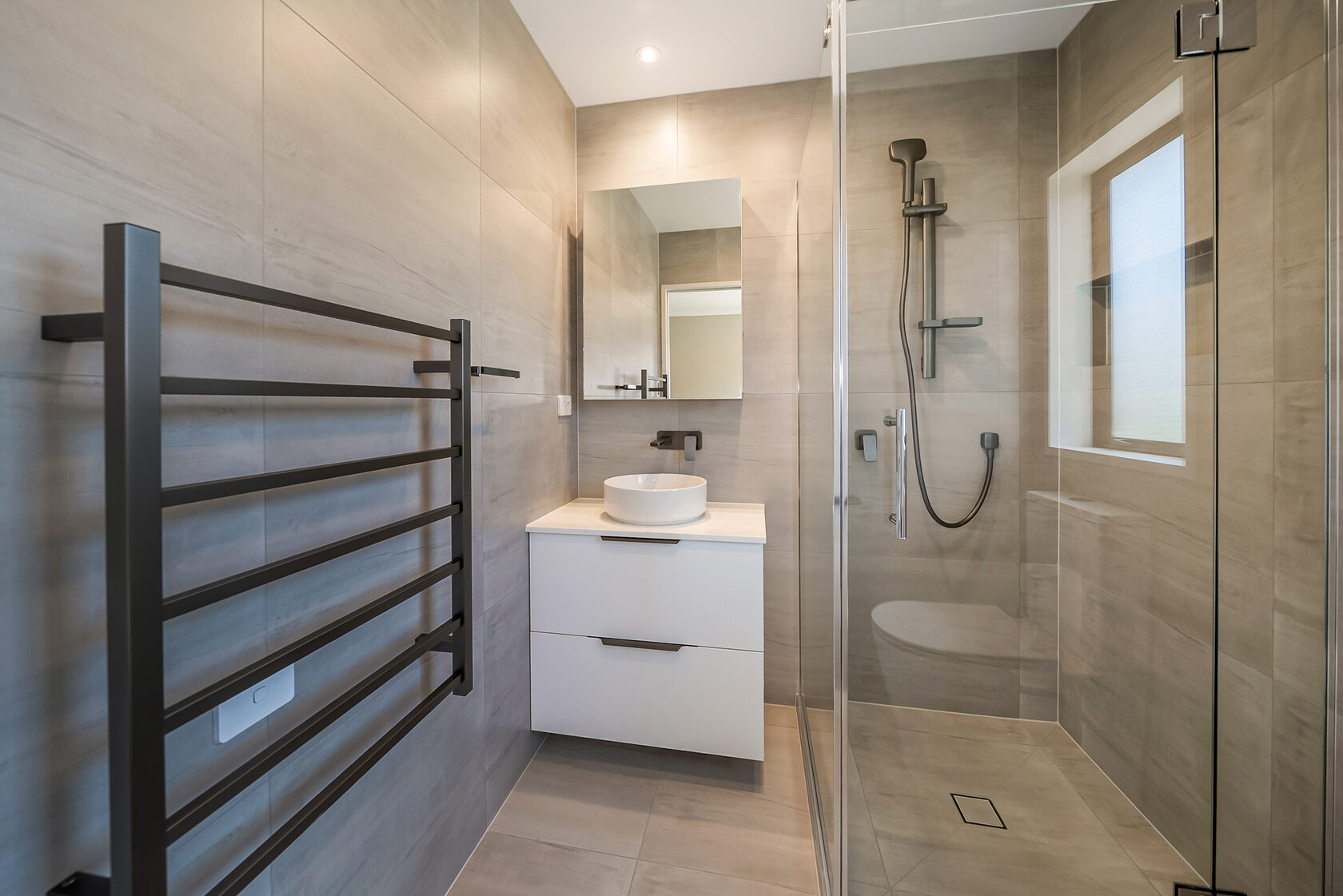 Flat Bush home renovation: Modern compact bathroom with beige wall tiles, floating white vanity with round vessel basin, black tapware, framed mirror, heated black towel rail, and a glass walk-in shower with rainfall and handheld showerhead.