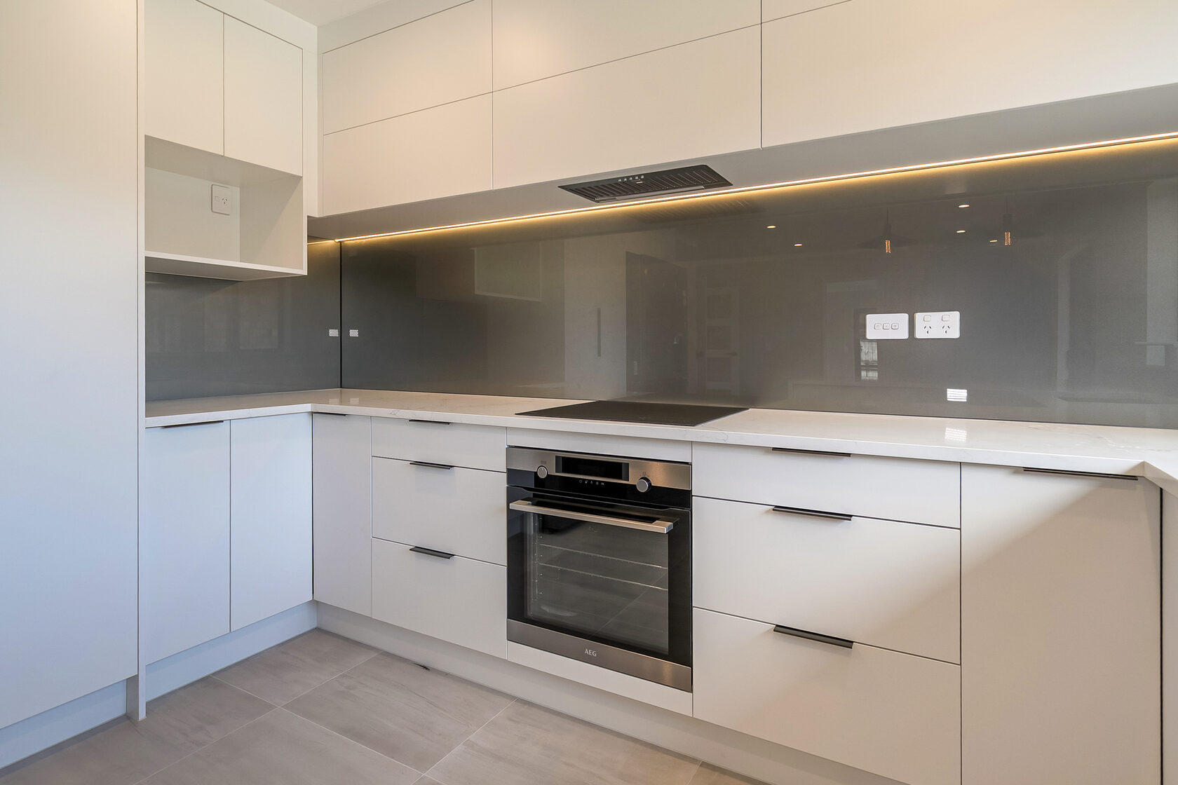 Modern white kitchen cabinetry with built-in oven and cooktop, grey glass splashback, under-cabinet lighting, and sleek black handles.