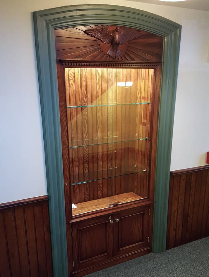 Historic church built-in display cabinet in stained wood with hand carved dove on top and lighted shelves 