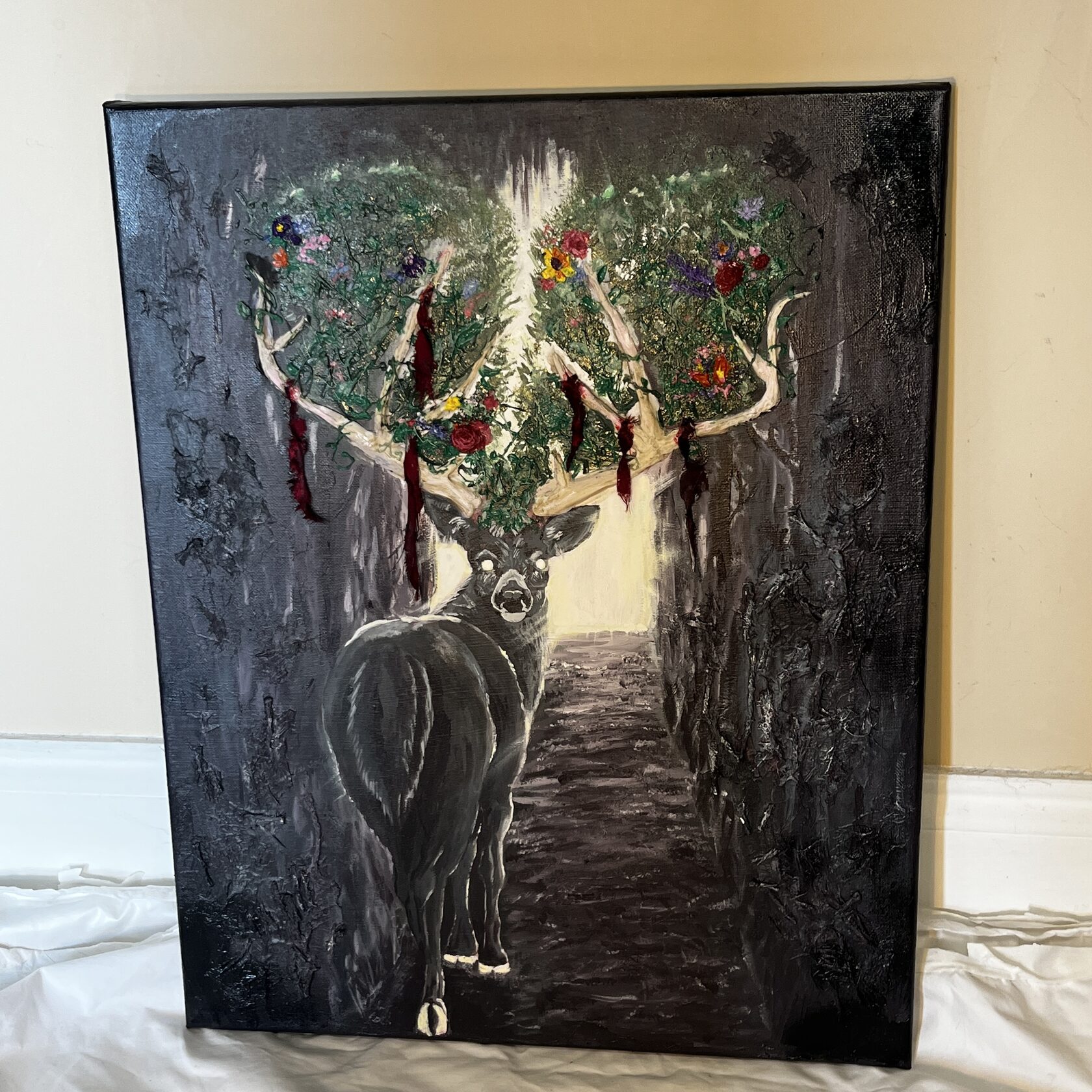 Come.: a buck turns toward the viewer on a forest path; flowers and fibers fill its antlers.
