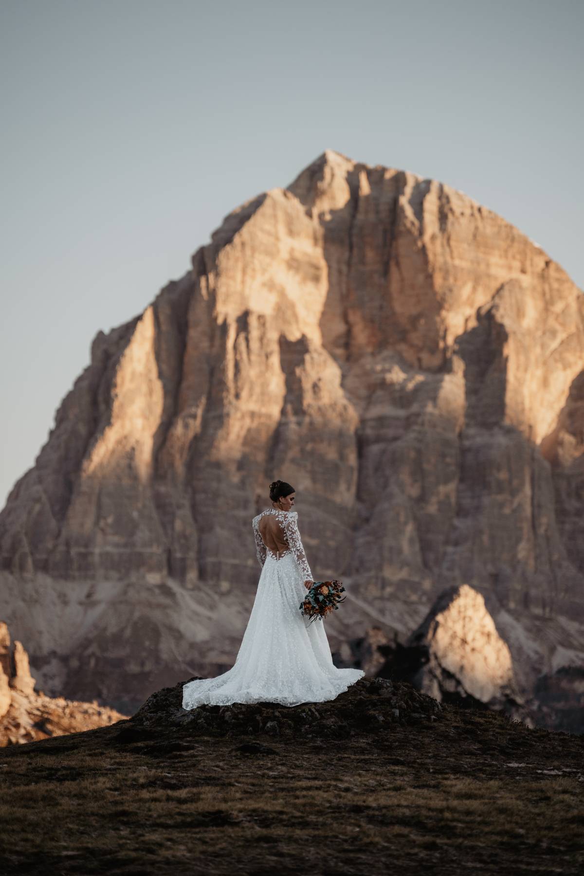 Bride in wedding dress looking toward a massive Dolomites rock face at sunrise, Italy, adventurous portrait