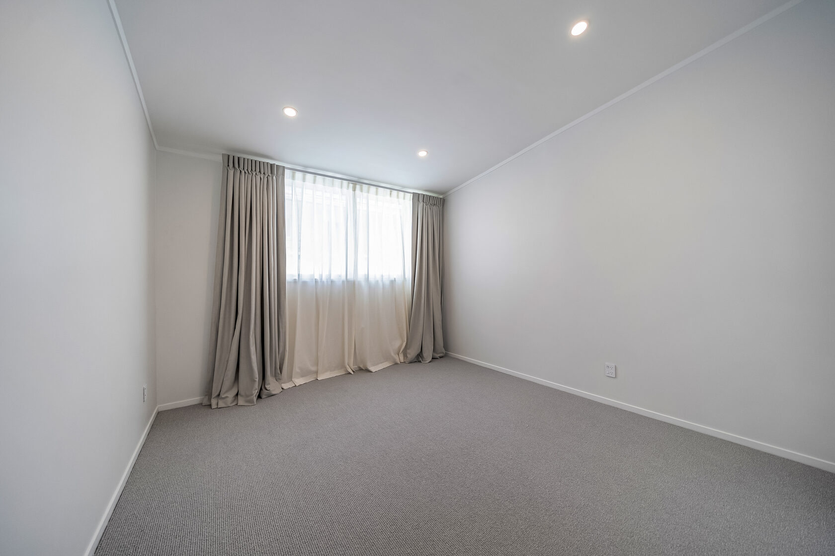 Minimalist bedroom with neutral walls, carpet flooring, recessed ceiling lights, and window with full-length curtains.