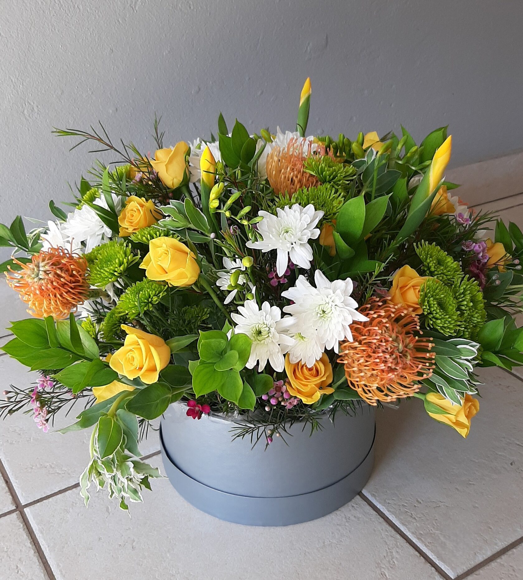 a vibrant floral arrangement in a round gray hat box, featuring yellow roses, white chrysanthemums, and orange leucospermum flowers