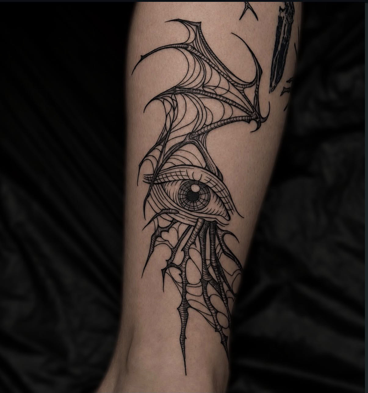 Tattoo of an eye with a textured iris, set in spiky web-like forms resembling wings above and branching roots below.