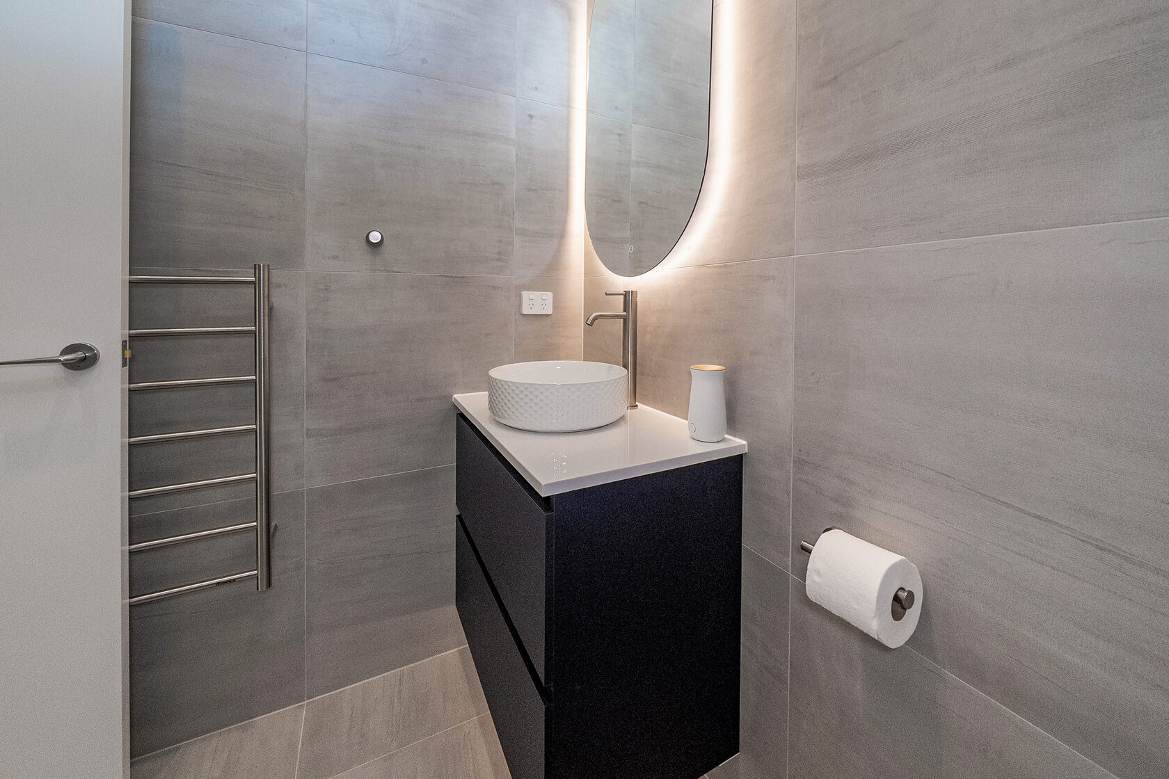 Contemporary bathroom with grey tiled walls, floating vanity, vessel sink, backlit mirror, and heated towel rail.