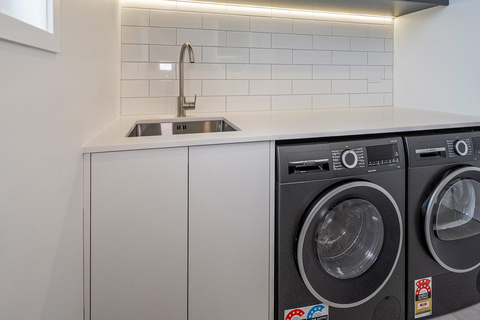 Modern laundry area with stainless steel sink, white subway tile splashback, and front-loading washing machine.