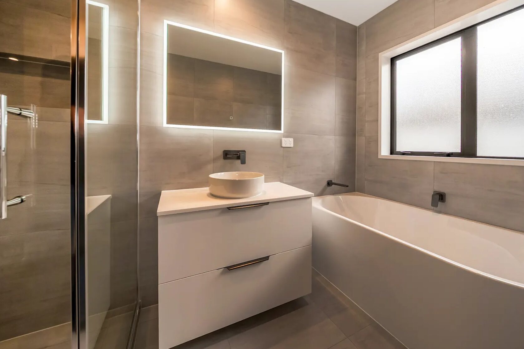 After: Renovated family bathroom with a glass shower, white vanity, illuminated mirror and bathtub