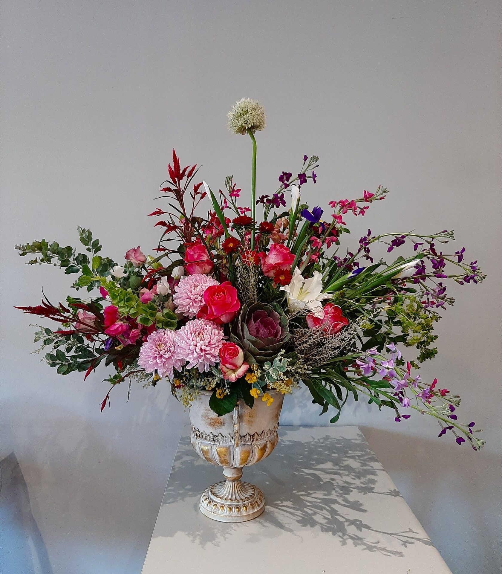 A luxurious floral arrangement in a classic antique urn, featuring peony-style roses, chrysanthemums, ornamental kale, and wildflowers