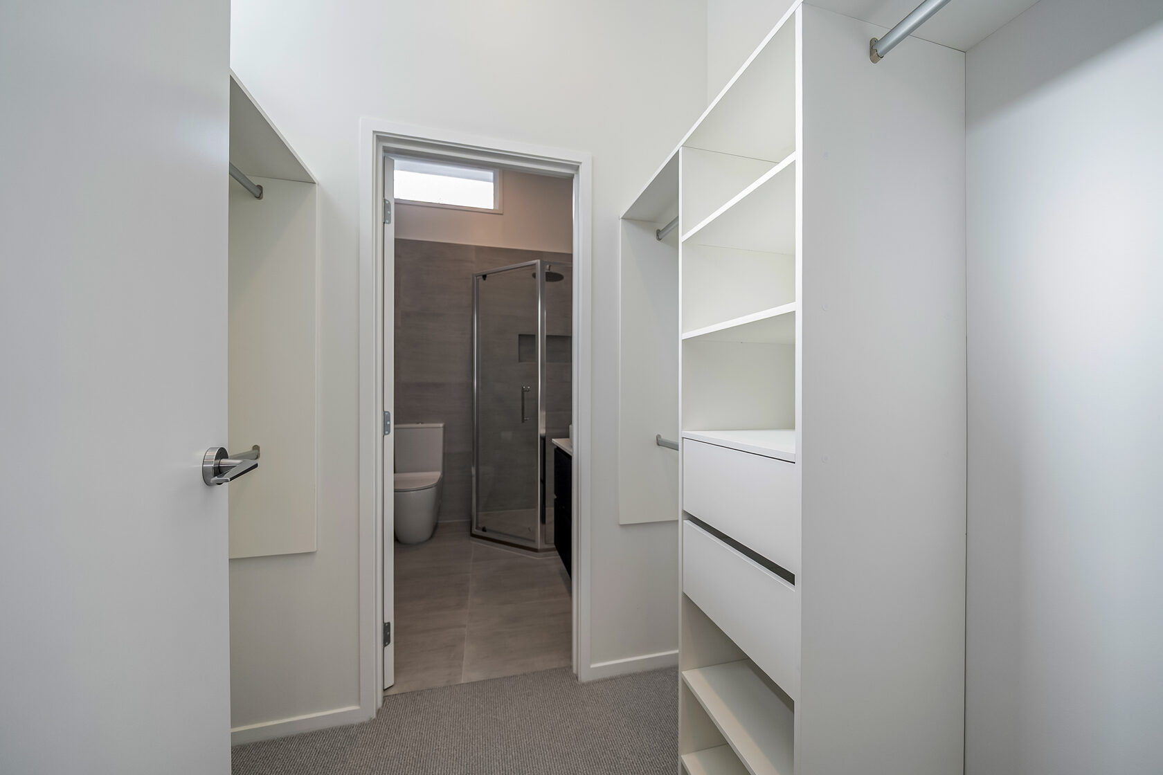 Walk-in wardrobe with built-in shelving and hanging rails leading to a modern ensuite bathroom with glass shower.