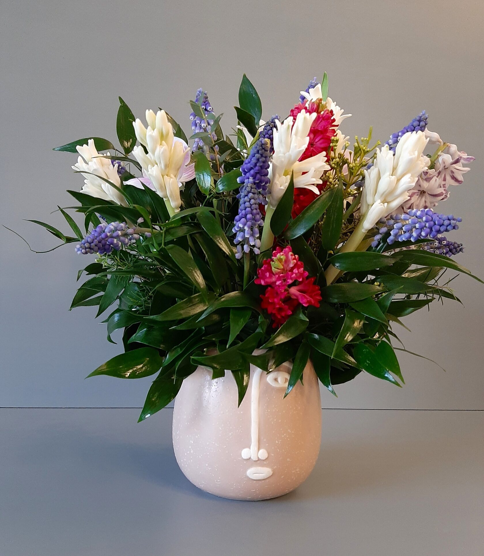 A beautiful bouquet of hyacinths, muscari, and greenery in a unique ceramic vase featuring a face.