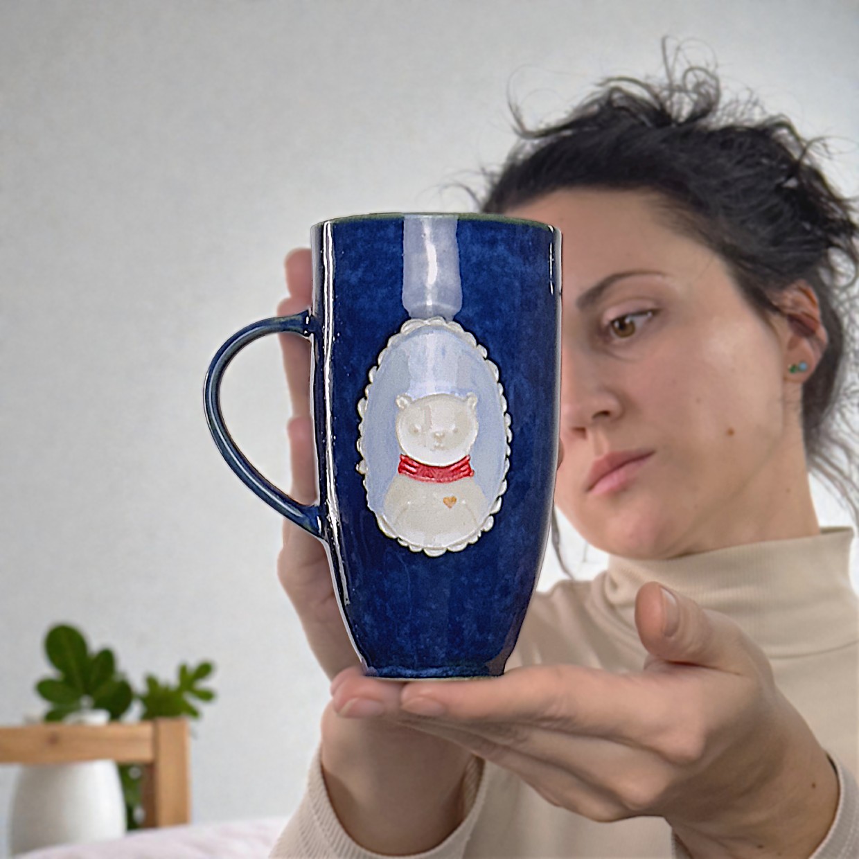 A woman holding a handmade blue ceramic mug featuring a teddy bear relief. Cozy and artistic atmosphere.