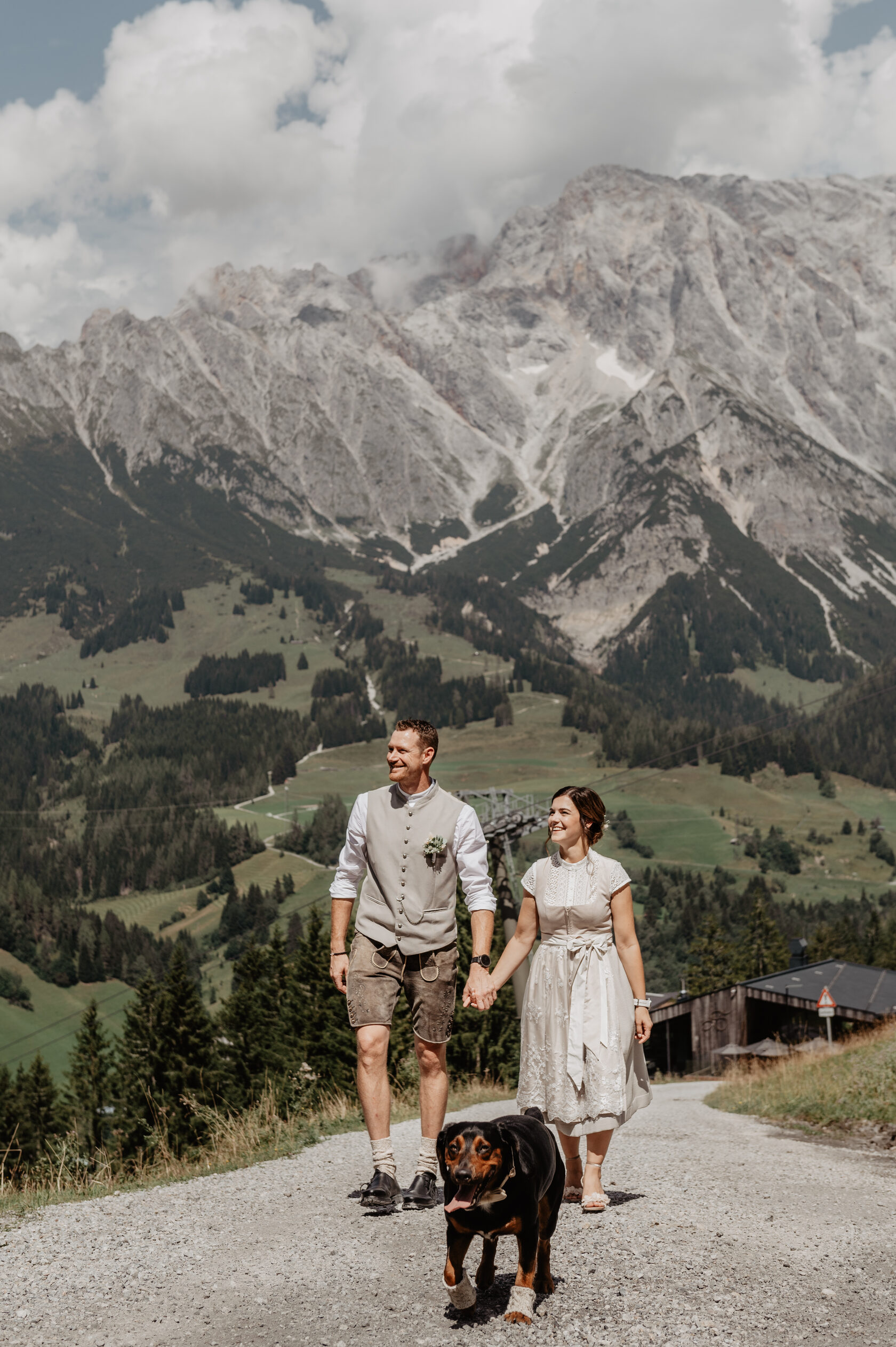 After‑wedding adventure in the Alps — couple walking with dog in traditional attire on mountain trail.