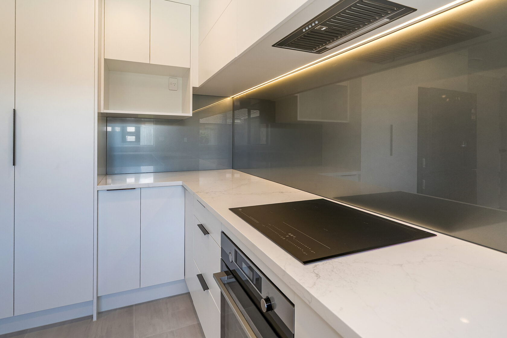 Close-up of modern kitchen corner with induction cooktop, built-in oven, grey glass splashback, white cabinetry, and warm under-cabinet LED lighting.