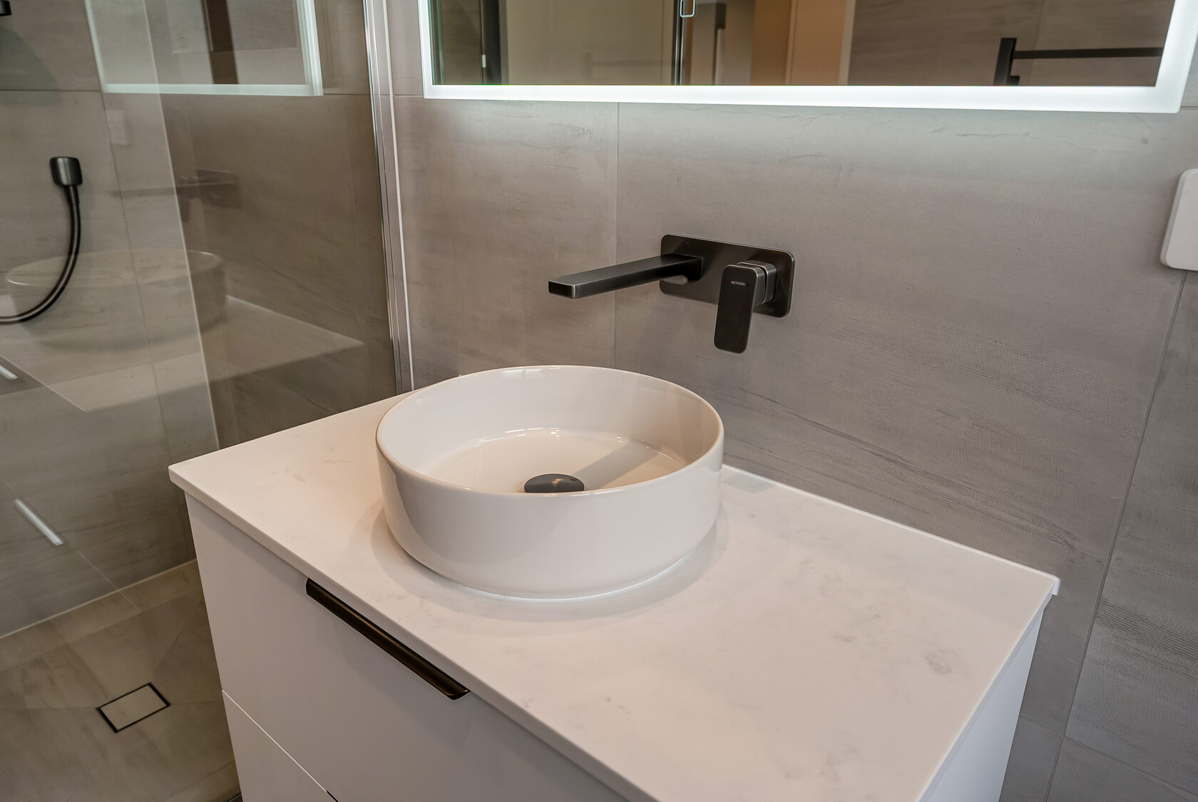 Close-up of a modern round white vessel basin on a stone-look countertop, paired with matte black wall-mounted tapware and minimalist floating vanity cabinetry.