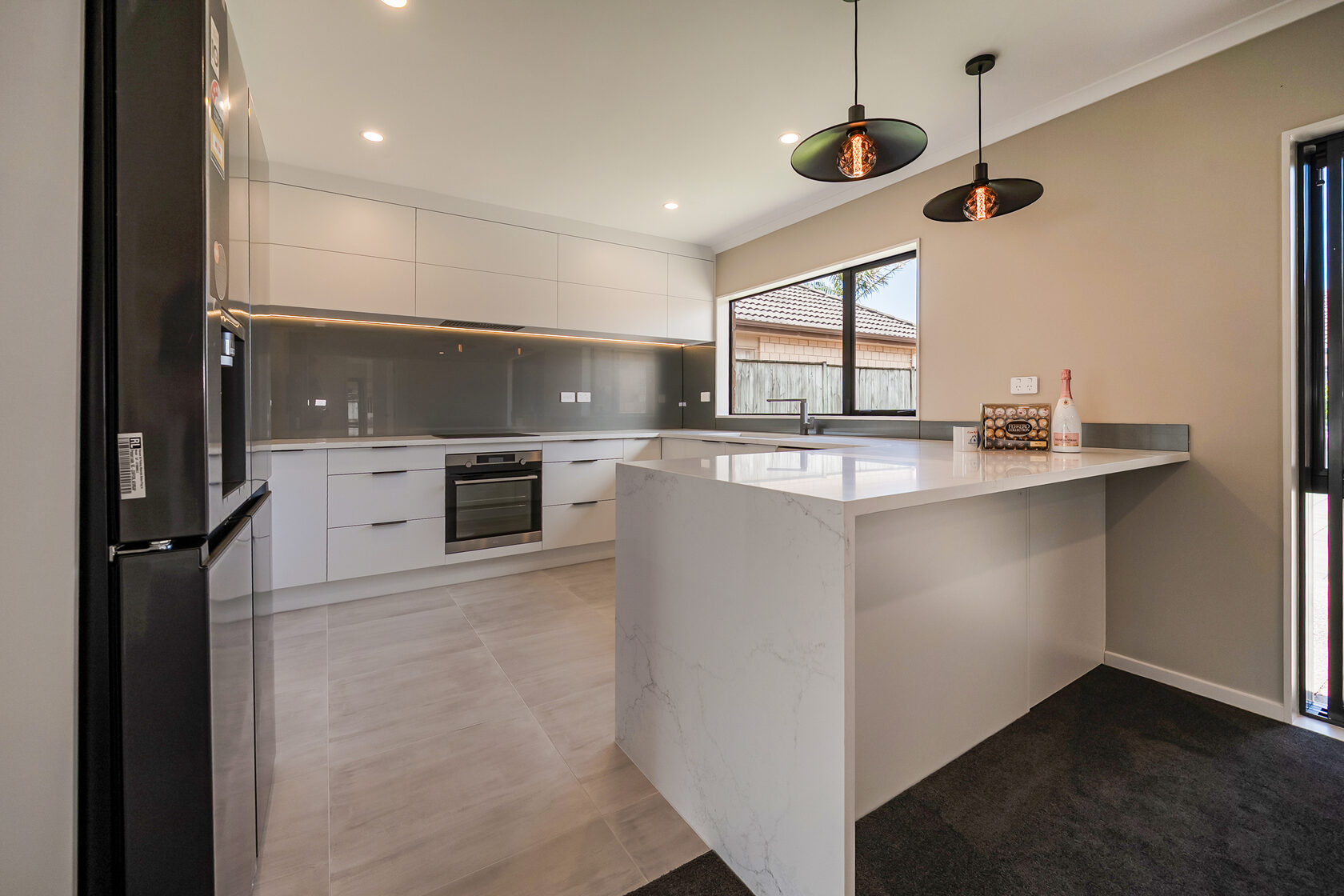 Spacious contemporary kitchen with large marble-look island, white handleless cabinetry, grey glass splashback, black fridge, and modern pendant lighting.