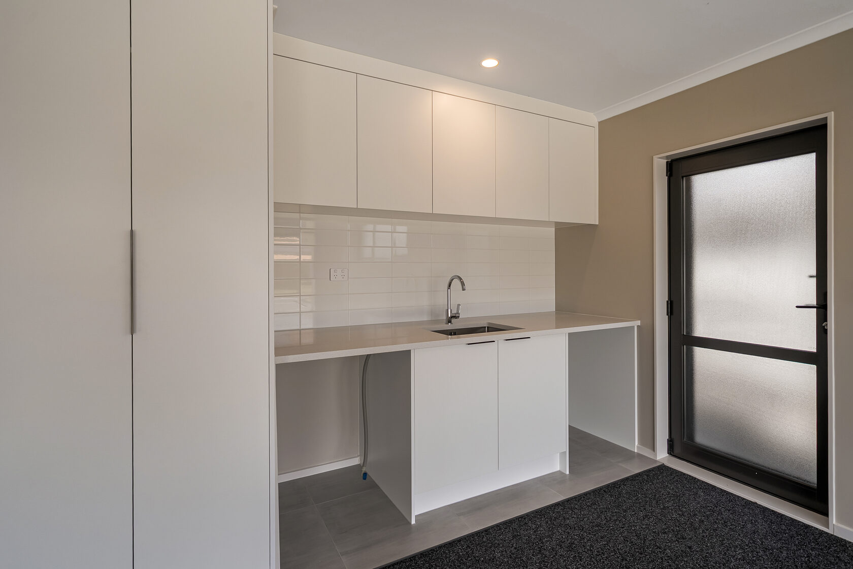 Compact laundry area with white overhead cabinets, tiled splashback, stainless steel sink, and frosted glass exterior door.