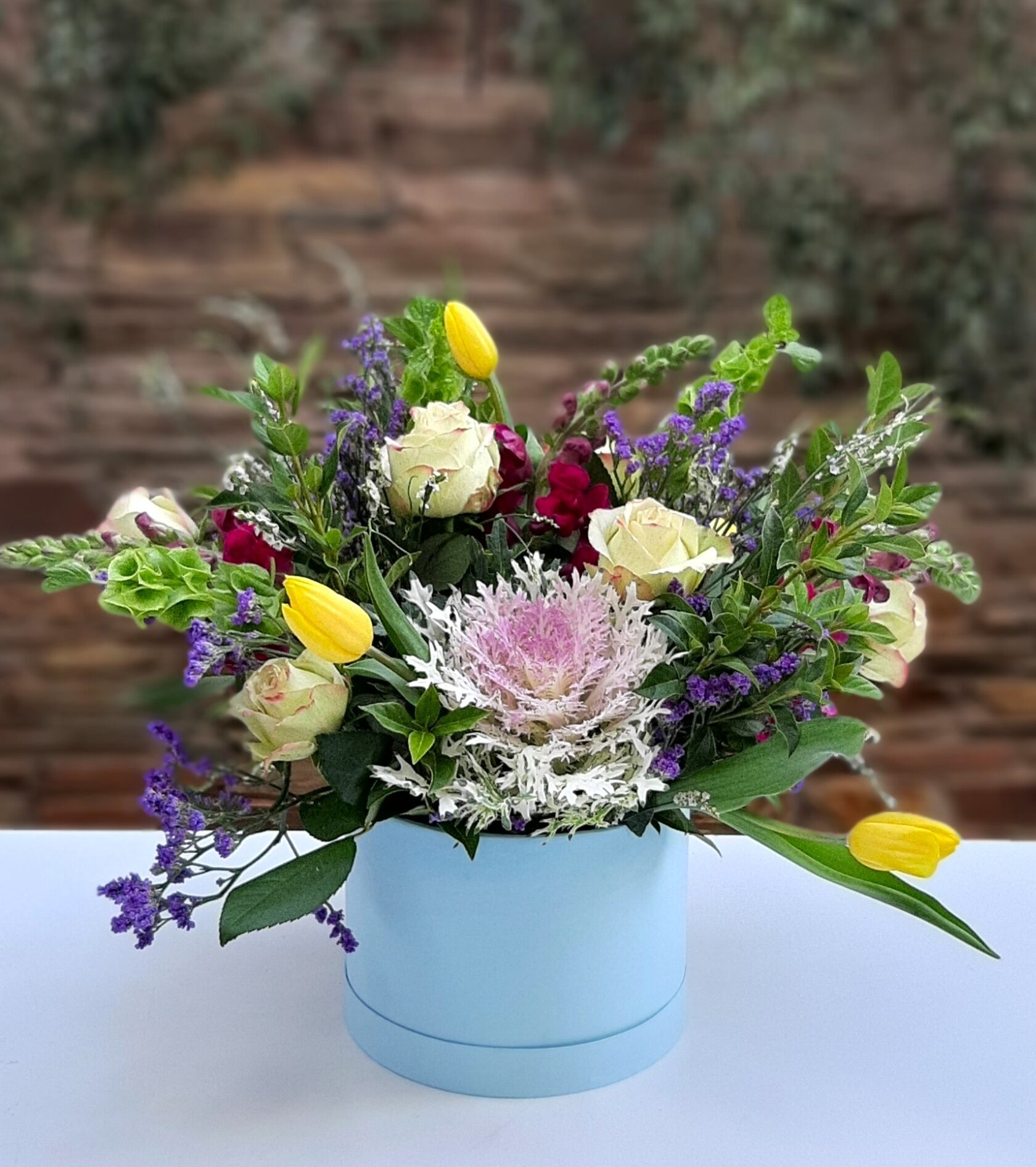 A delicate floral arrangement in a round blue hat box, featuring ornamental cabbage (Brassica), light-colored roses, yellow tulips, and purple statice.