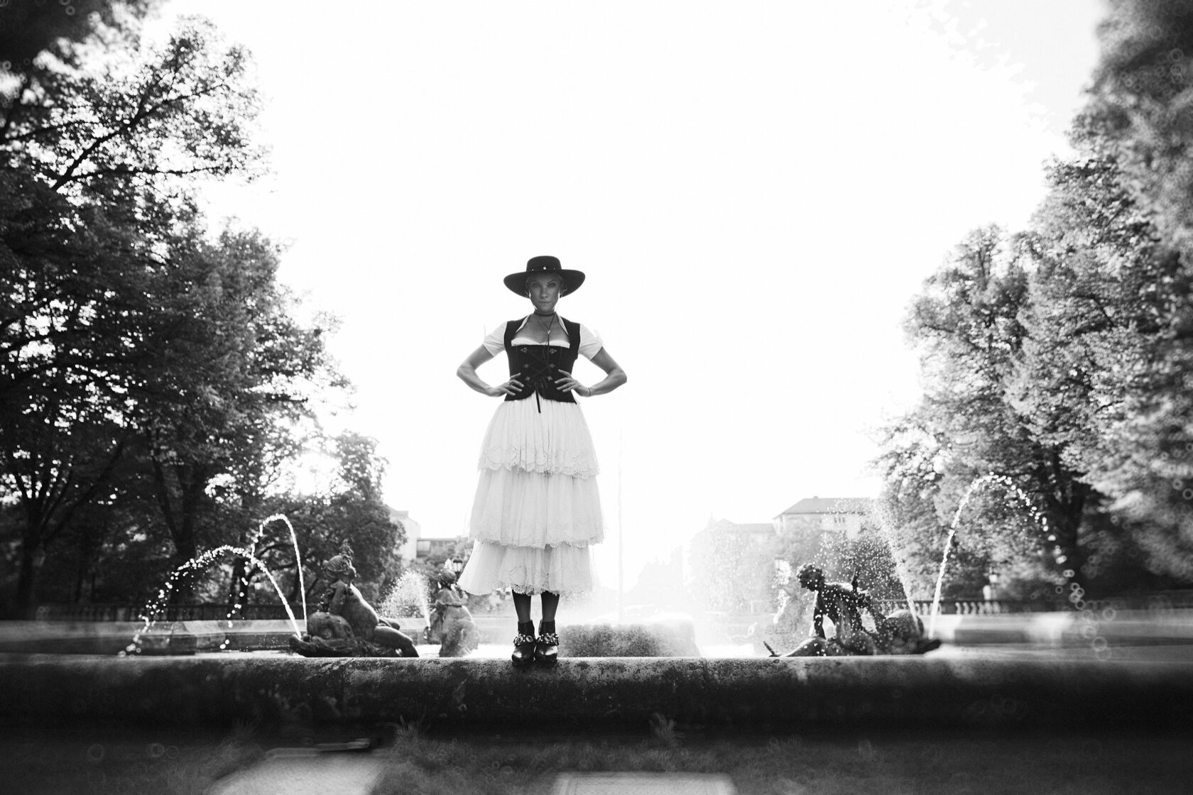 Schwarz-Weiß-Portrait einer Frau im Dirndl mit Hut vor einem Brunnen