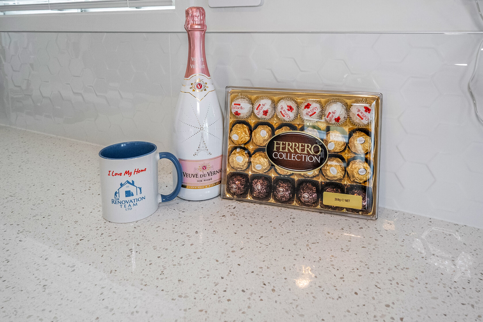 Luxury gift display with sparkling wine, Ferrero Collection chocolates, and branded mug on modern kitchen countertop.