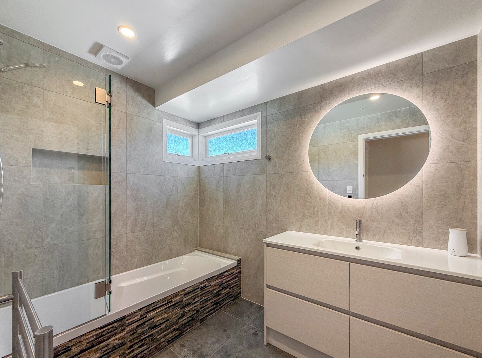 Renovated bathroom with a shower over the bath, grey wall tiles and an illuminated round mirror
