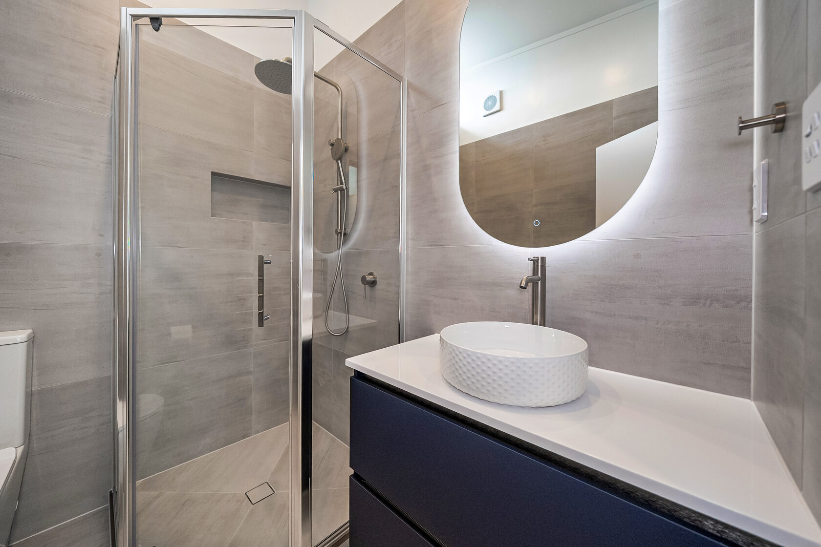 Modern bathroom with glass shower, textured vessel sink, floating vanity, and backlit oval mirror.