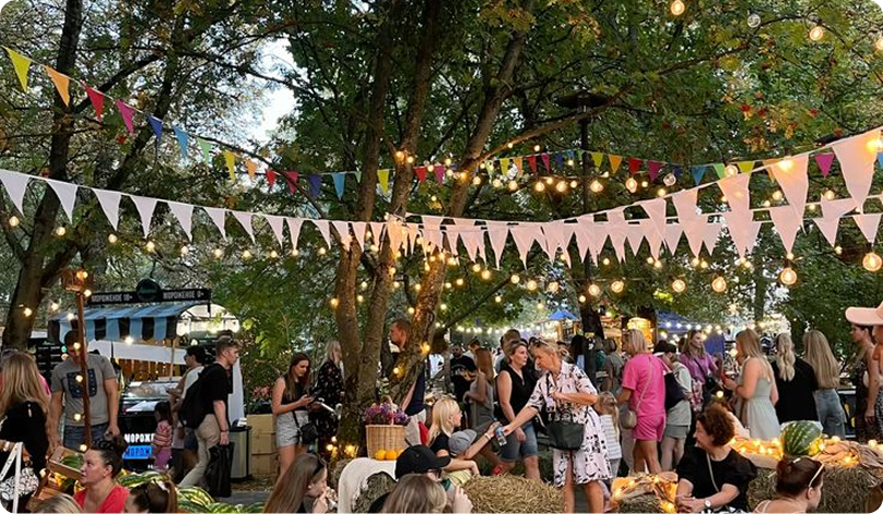 Udoban letnji korporativ u formatu uličnog festivala na otvorenom. Gosti u casual odeći opuštaju se na zelenom travnjaku, druže se u blizini terena za hranu i fotozona. Drveće je ukrašeno vijencima i obojenim zastavicama, stvarajući opuštenu prazničnu atmosferu. Jasan primer neformalnog formata za organizovanje korporativnih događaja na otvorenom.