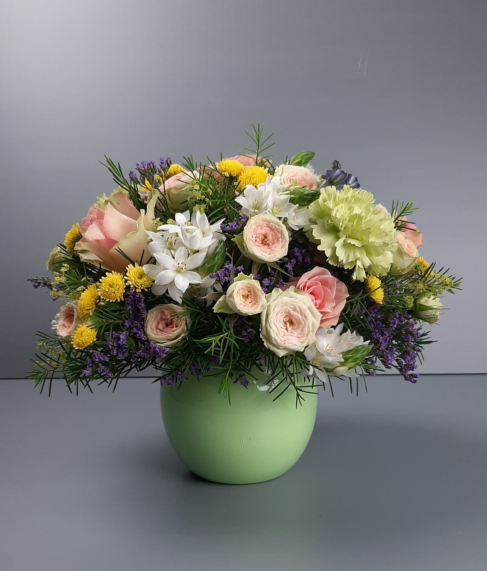 A round floral arrangement in a light-green round planter, featuring delicate pink and cream spray roses, small white and yellow flowers, and greenery.