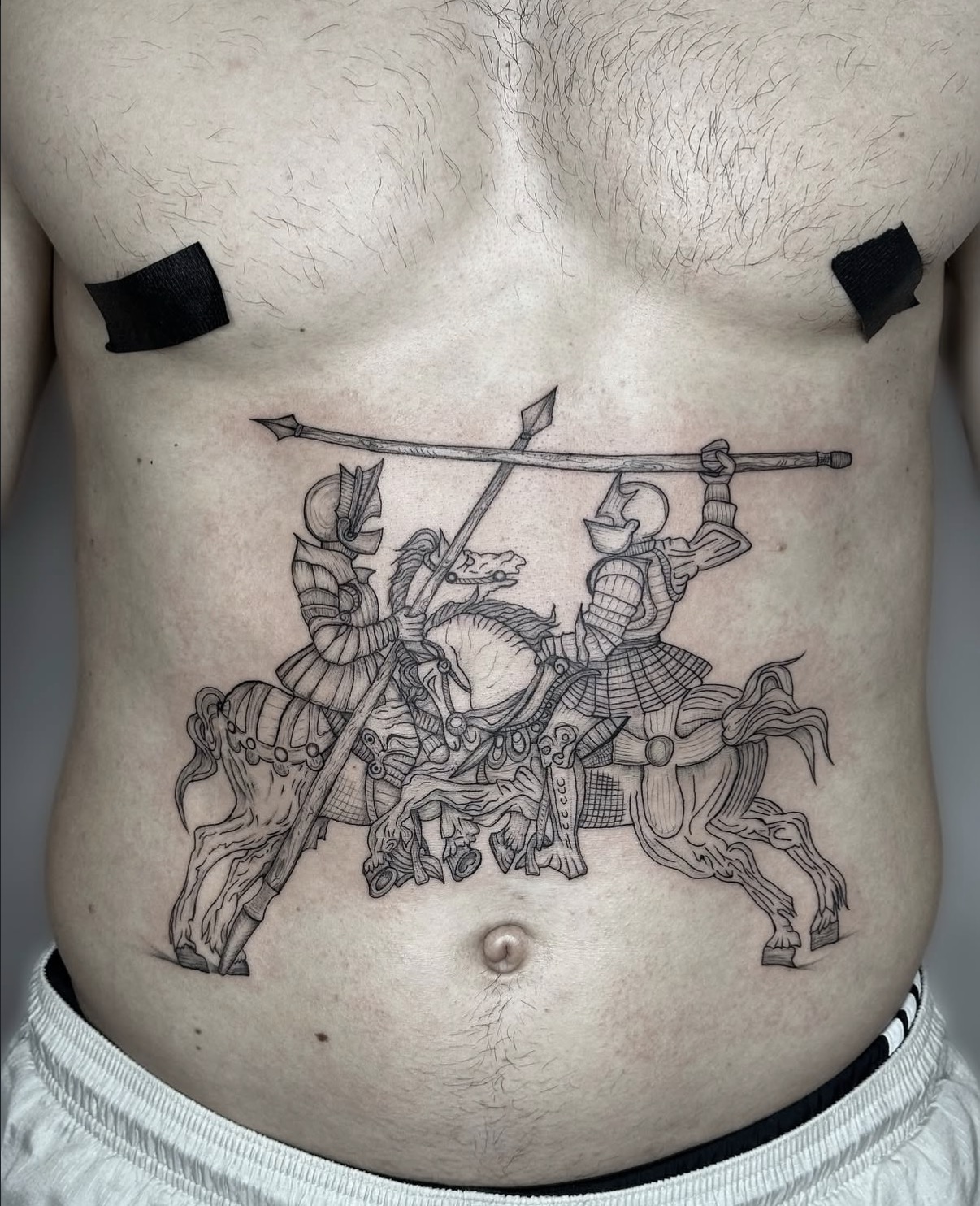 Abdomen tattoo of two armoured knights on horseback with crossing lances, helmets and decorated horse tack.