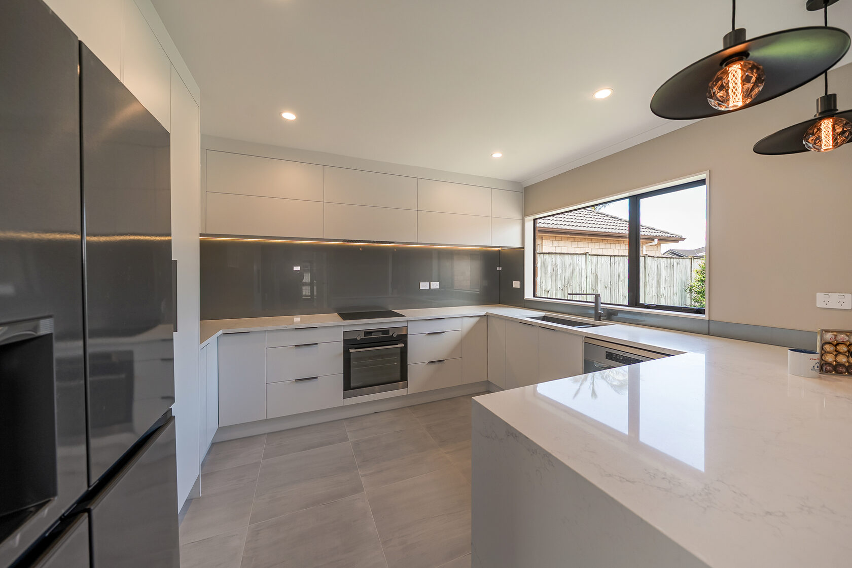 Contemporary white kitchen with handleless cabinetry, grey glass splashback, built-in oven and cooktop, large window, and spacious island with stone countertop.
