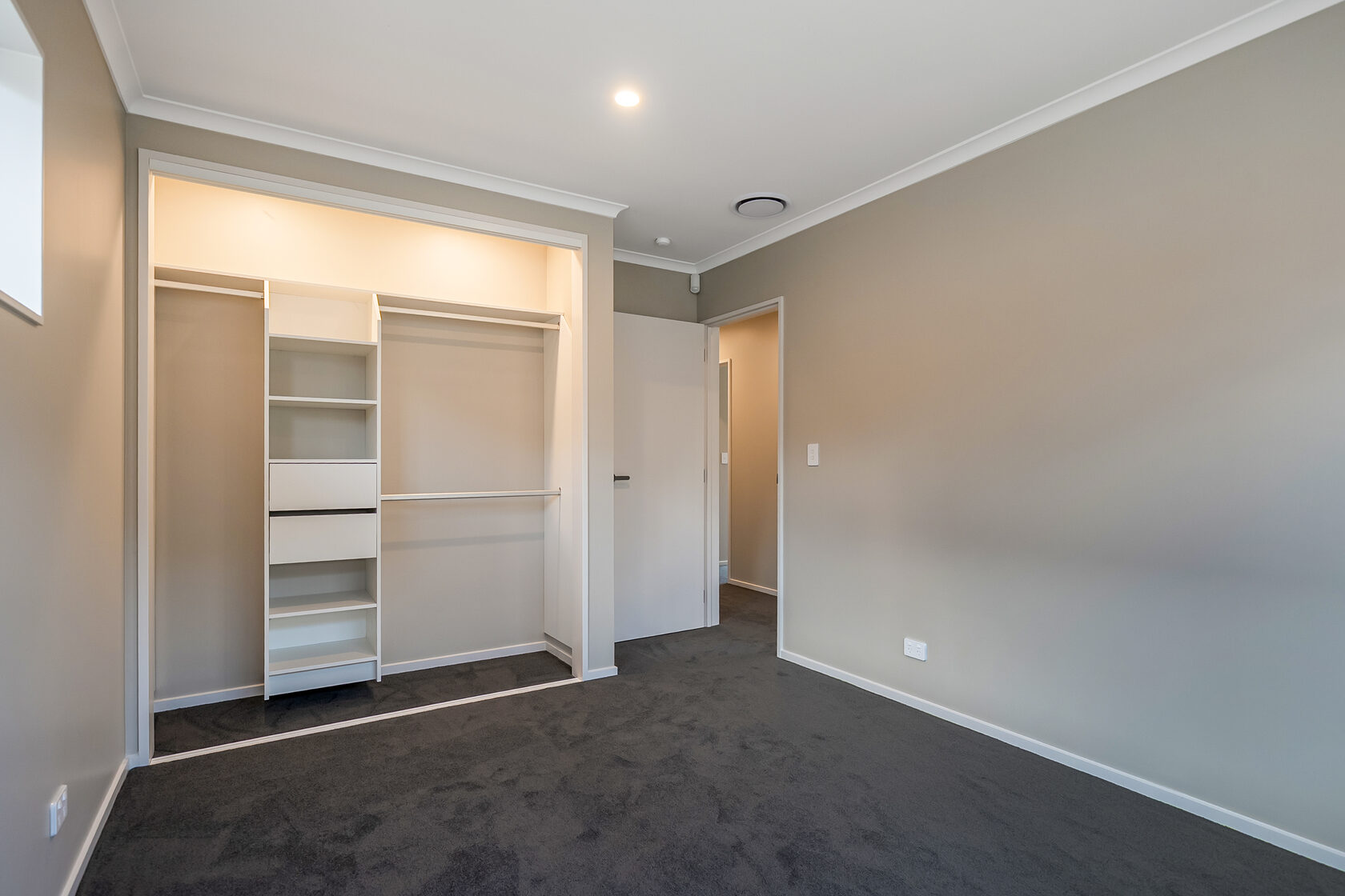 Empty bedroom with dark carpet, neutral walls, recessed lighting, and an open built-in wardrobe with shelving and hanging space.