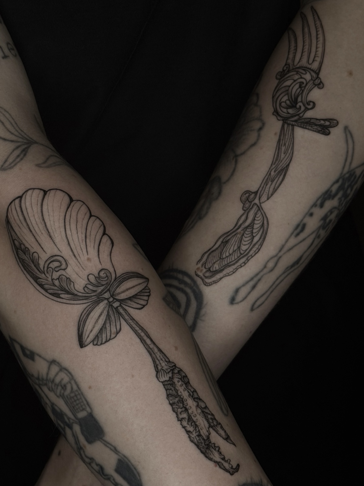 Two forearms with tattoos of a spoon with a shell-shaped bowl and a fork with scrollwork, small wings and textured handles.