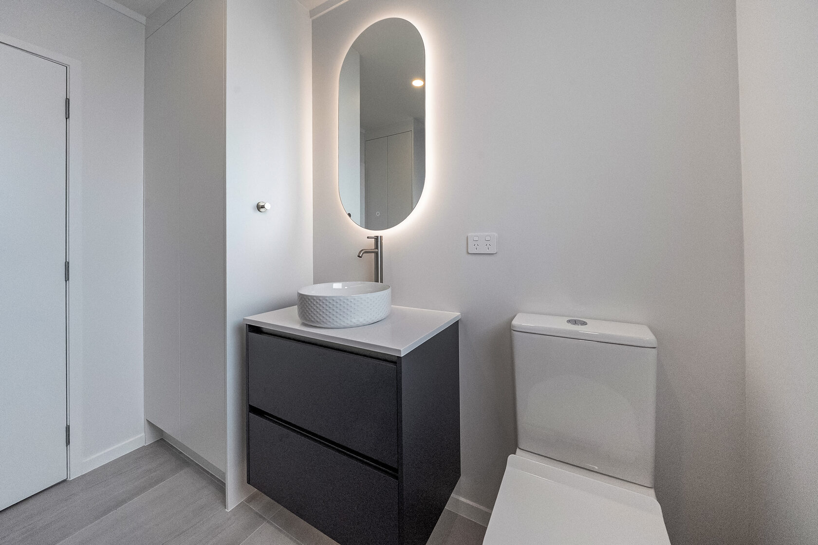 Compact modern bathroom with floating vanity, vessel sink, oval backlit mirror, and minimalist toilet.