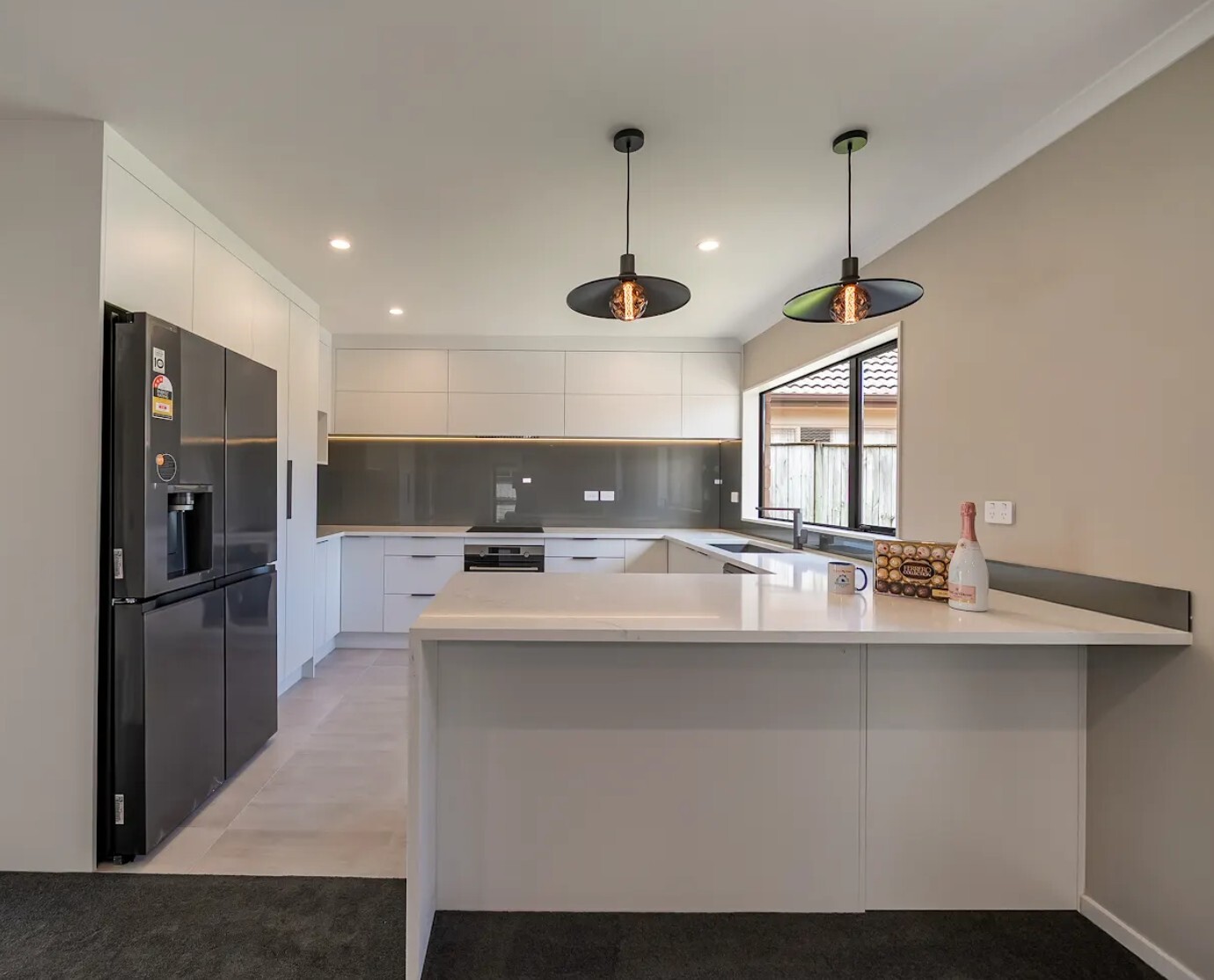 After: Renovated Flat Bush kitchen with a white peninsula, grey splashback, black refrigerator and pendant lights